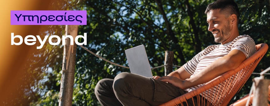 A man in a striped shirt smiles while using a laptop in an orange chair outdoors. Sunlight filters through trees. Text reads "Υπηρεσίες beyond."